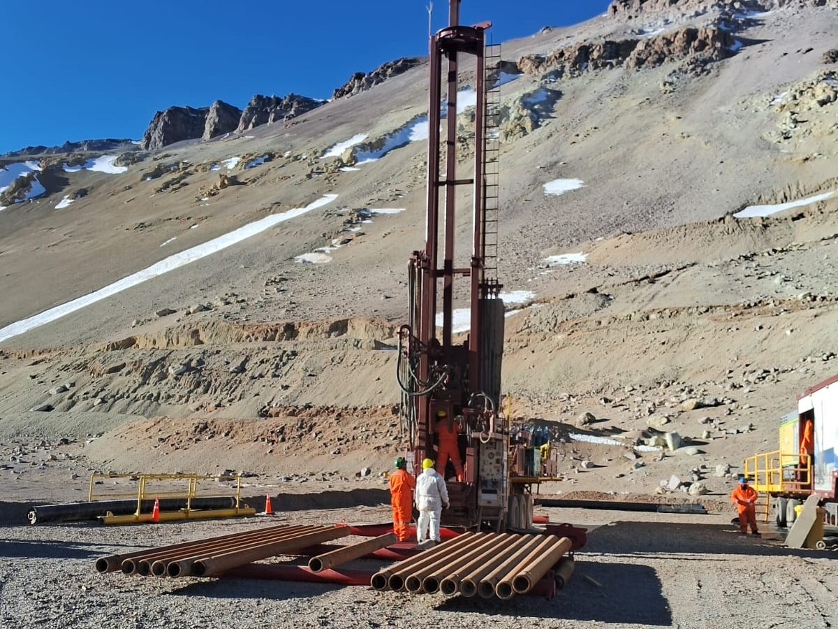 Equipo en plataforma al pie de una ladera con nieve, con barras de perforación tendidas en el sitio / Rig on its pad at the foot of a snow-patched slope, with drill rods laid out on site