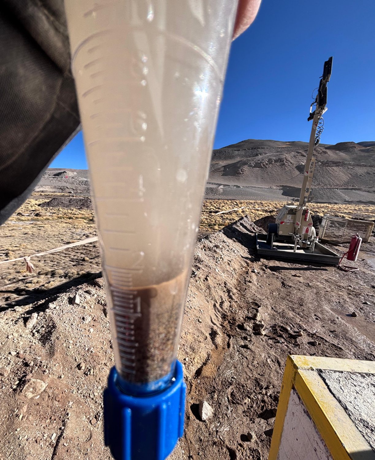 Muestra de agua del pozo en probeta graduada, con el equipo de perforación al fondo / Well water sample in a graduated cylinder, with the rig behind